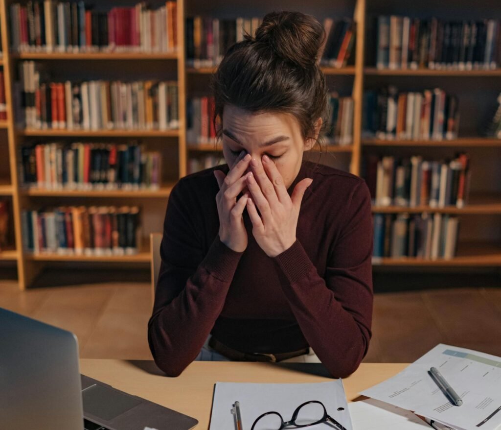 Woman feeling stressed while studying in a library, surrounded by books and laptop.