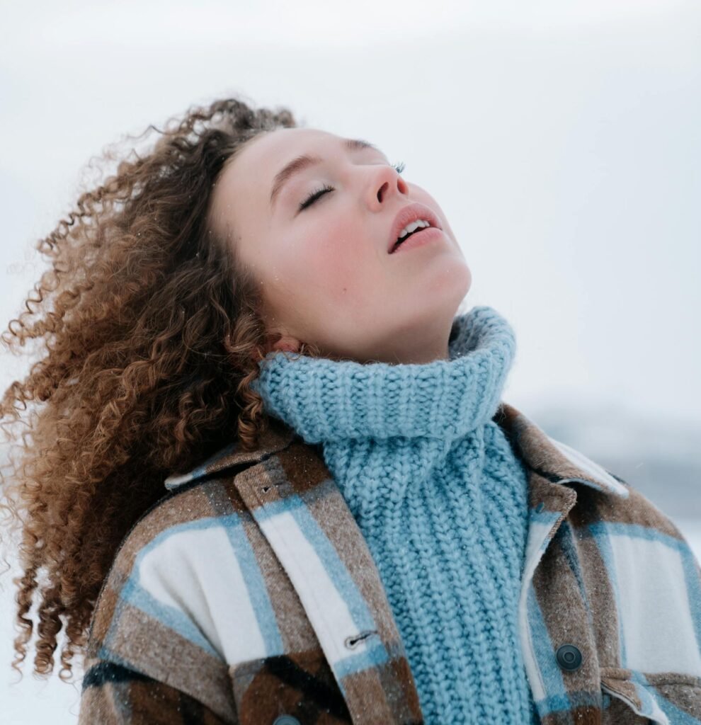 Woman with curly hair in winter attire standing outdoors with eyes closed, expressing serenity.