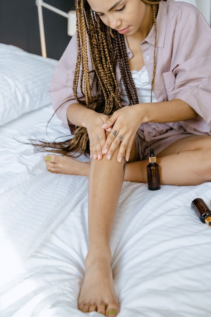 African American woman applying essential oil on leg indoors, focusing on skincare and relaxation.