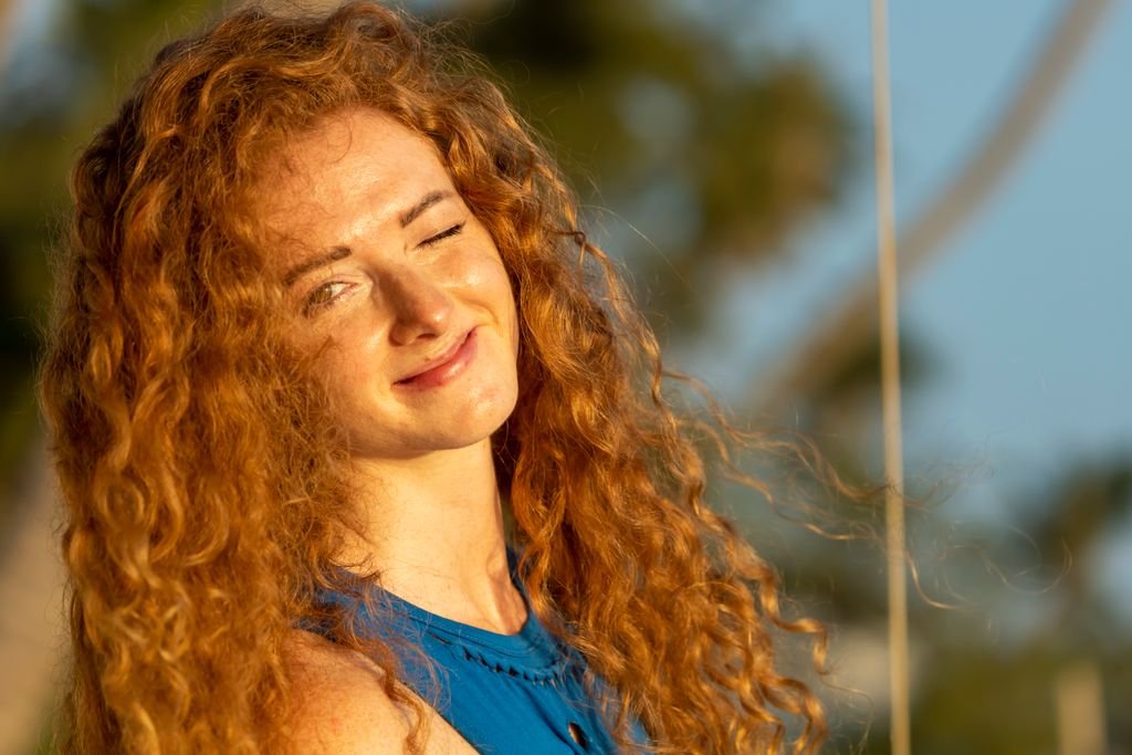 A cheerful woman with curly hair winking and smiling in sunlight.