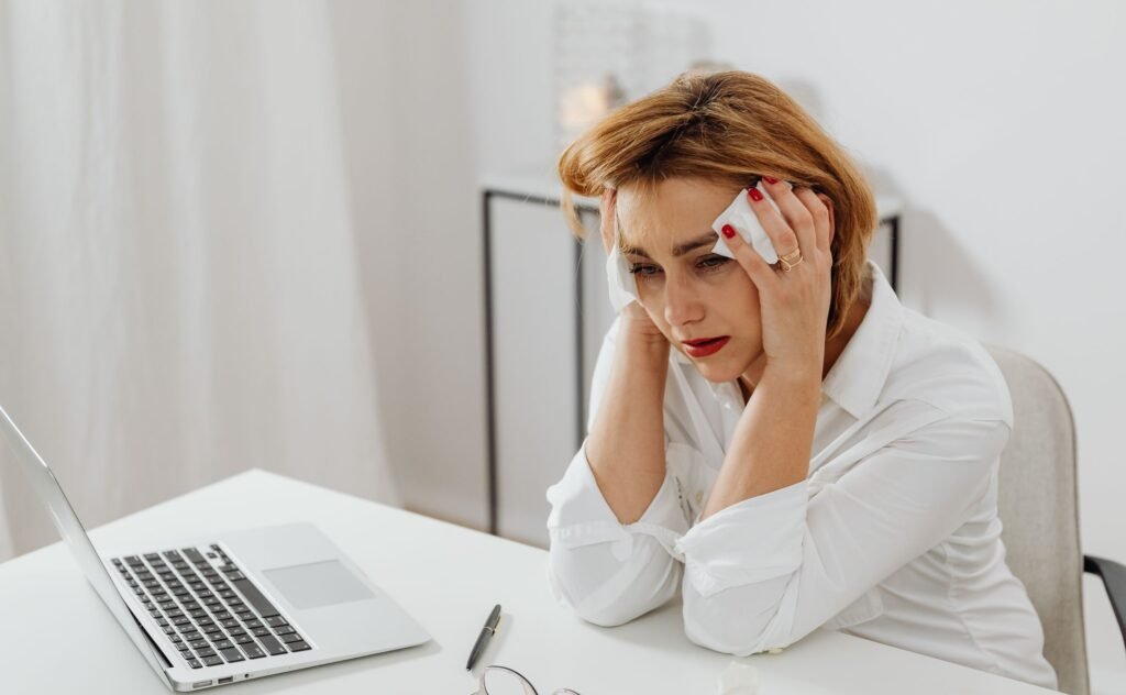 A woman in distress at her office desk, surrounded by tissues and a laptop.