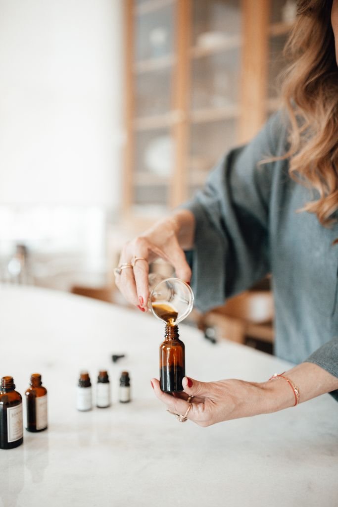 Close-up of a woman pouring essential oil into a glass bottle on a marble countertop.