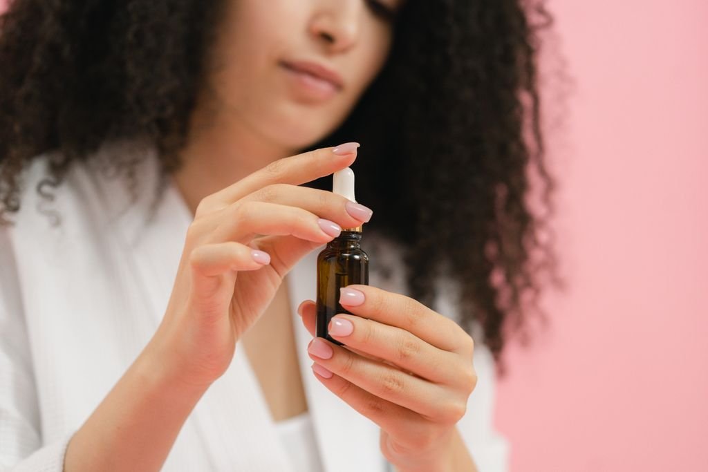 Close-up of woman holding a glass dropper bottle for skincare against a pink background.
