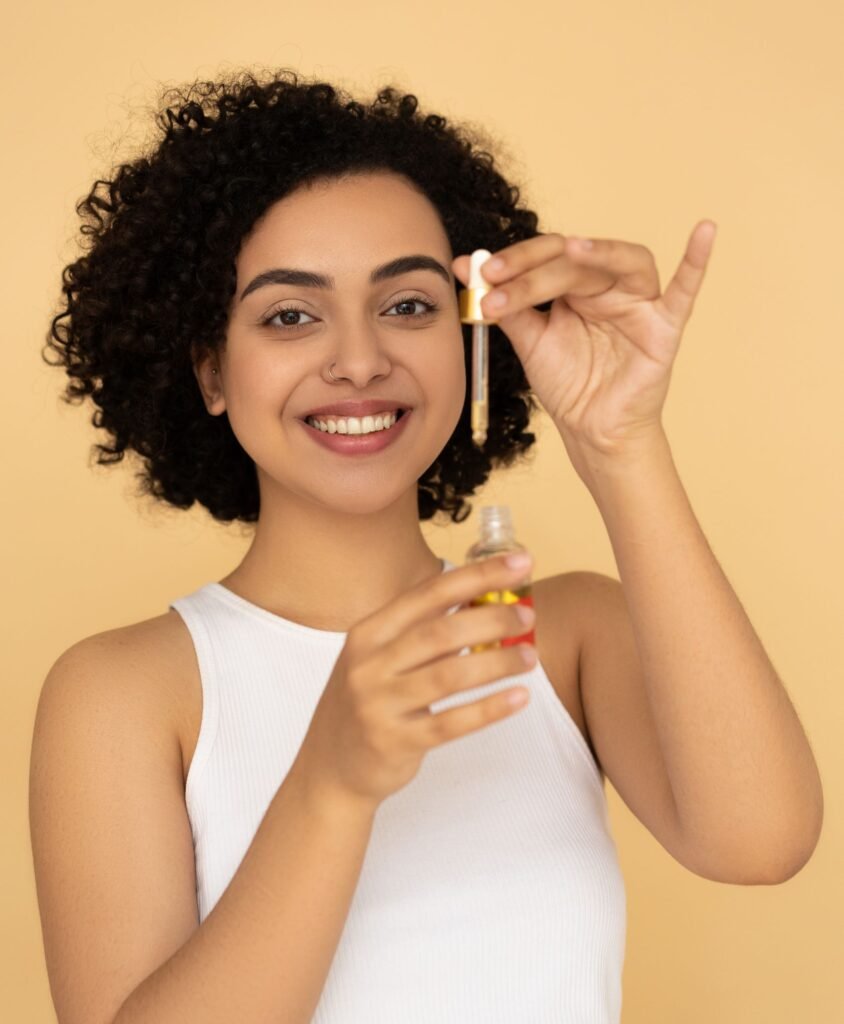 Smiling woman with curly hair applying skincare oil, promoting healthy skin and beauty care.