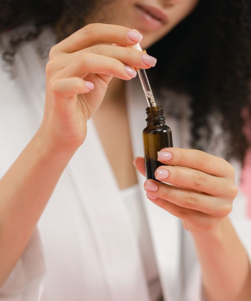 A woman in a white robe uses a dropper to apply skincare serum from a glass bottle.